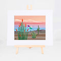 Hummingbird, Brenda's painting of a hummingbird among blooming ocotillo, prickly pear, and saguaros under a pink and blue sky, shown as a matted 8x10 art print