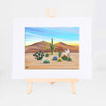 Desert in Bloom, Brenda's painting of saguaros, flowering prickly pear, ocotillo, agave, and wildflowers in warm sunlight, shown as a matted 8x10 art print
