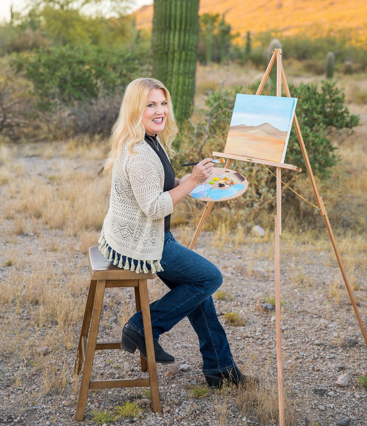 Brenda seated on a stool in the Arizona desert, in front of a canvas on an easel